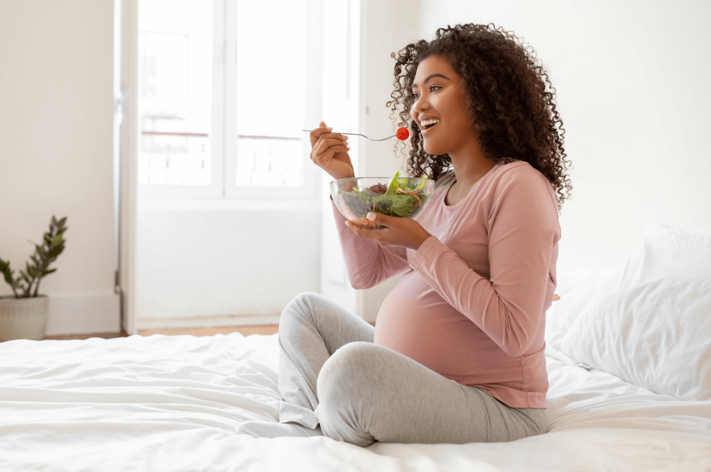 Embarazada sentada en el piso de su sala come ensalada verde. Prácticas de alimentación durante el embarazo.